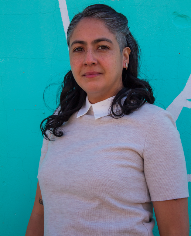 A person with long dark hair wearing a grey short-sleeve sweater and a white collar shirt stands in front of an aqua colored wall that has crisscrossing white diagonal lines.