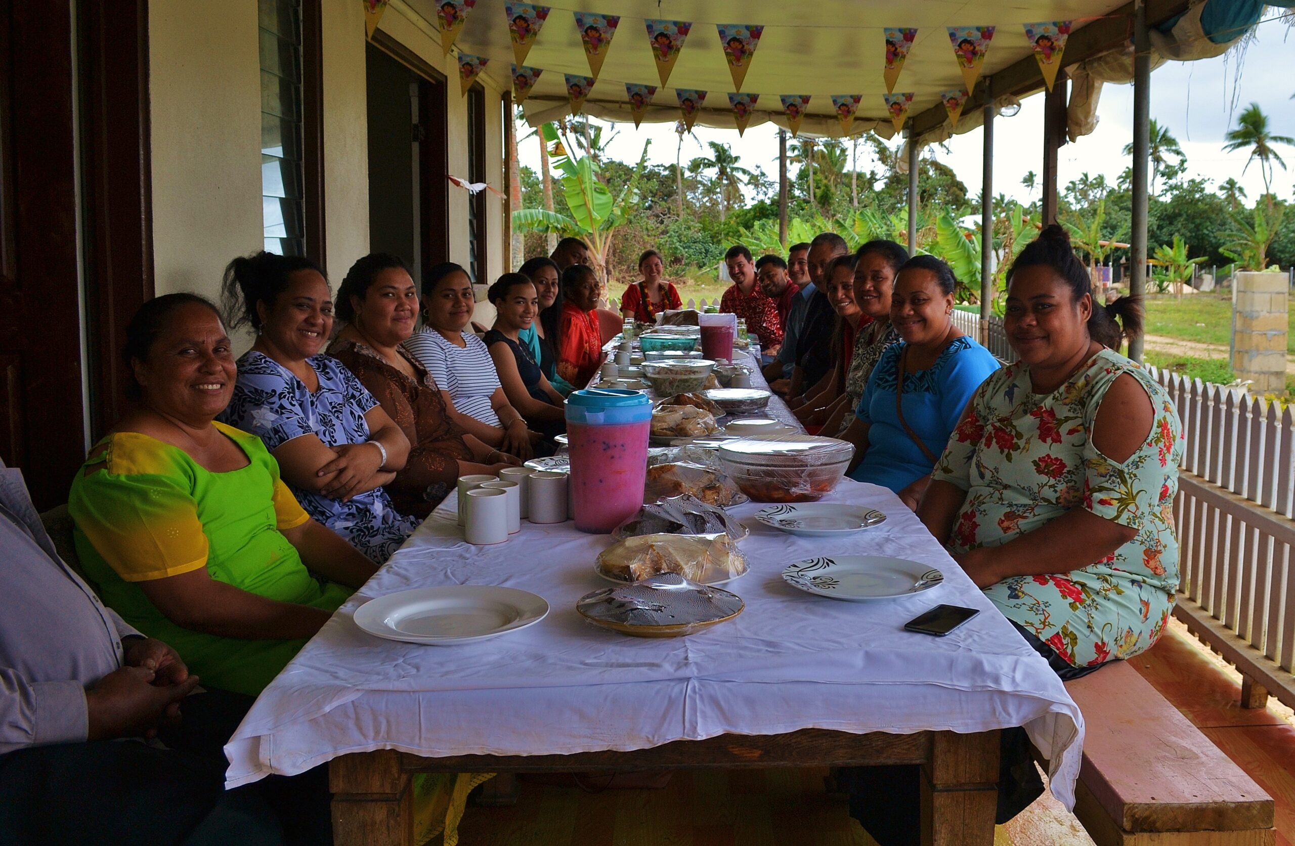 A large group of people sitting at a long outdoor table covered with a white table cloth and bowls of food and plates. Peaceworkers