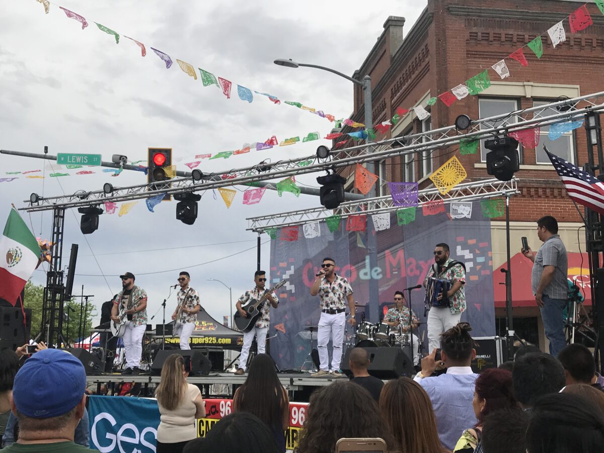 A musical band of six people stand by mics and hold three guitars and an accordion while on a stage singing to a crowd of people on a street corner. A Mexican flag, A U.S. flag, and small colorful flags hang from stage scaffolding. Immigrant.