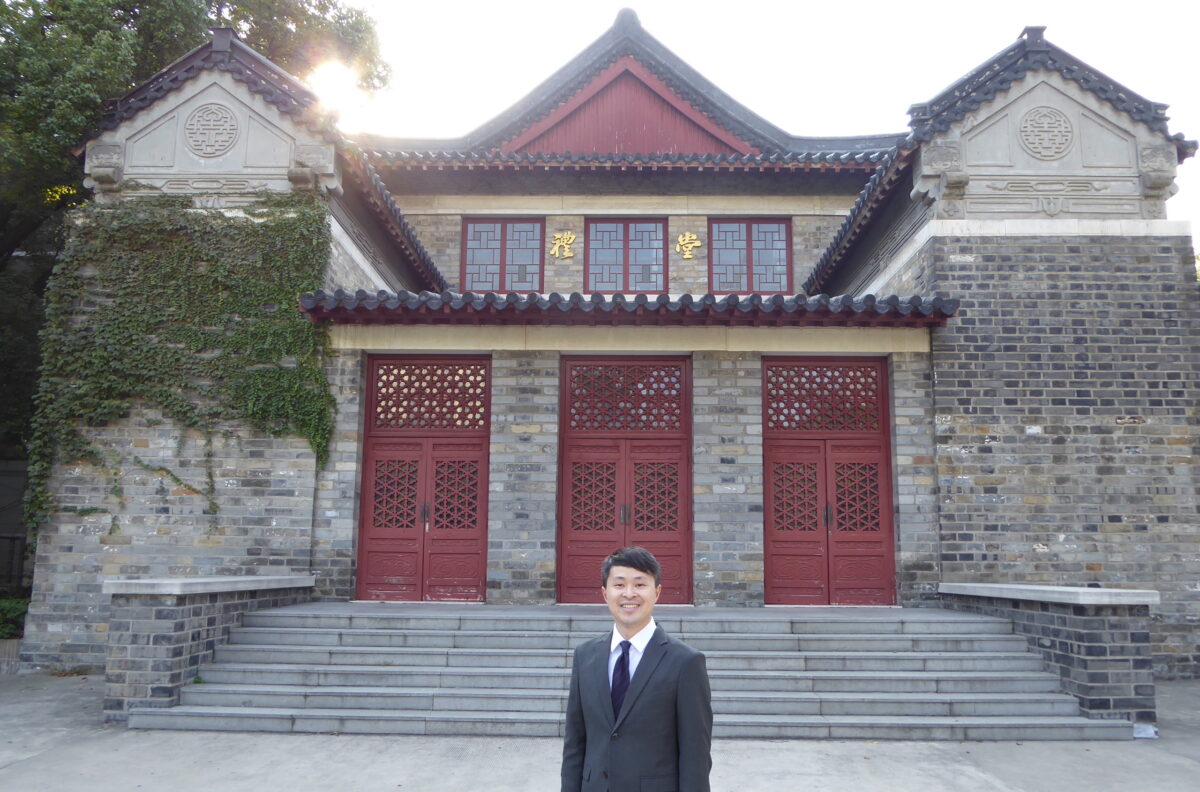 A person standing in front of a Nanjing University’s auditorium, a grey brick building with grey stone steps leading to three red wooden doors.