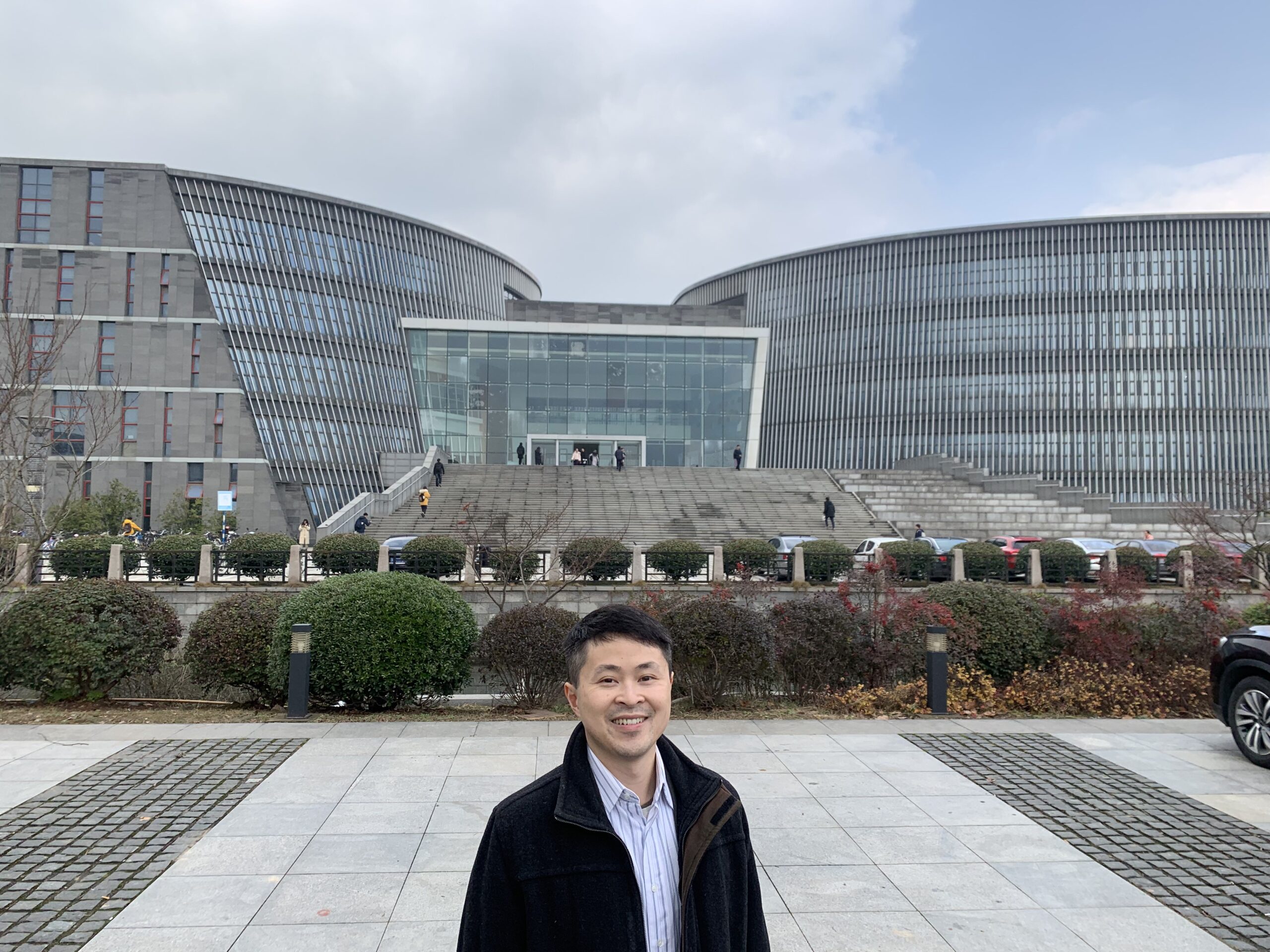 A person wearing a black jacket and a collar shirt stands in front of Nanjing University’s main library, a large modern building with sloping walls.