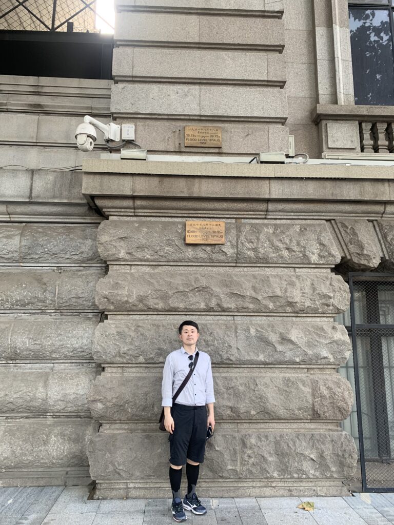 A person wearing a grey collar shirt and black shorts, socks, and sneakers stand in front of a large stone building underneath a plaque.