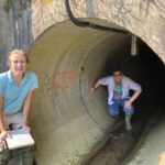 two researchers, one just outside and one inside a large underground pipe several feet in diameter