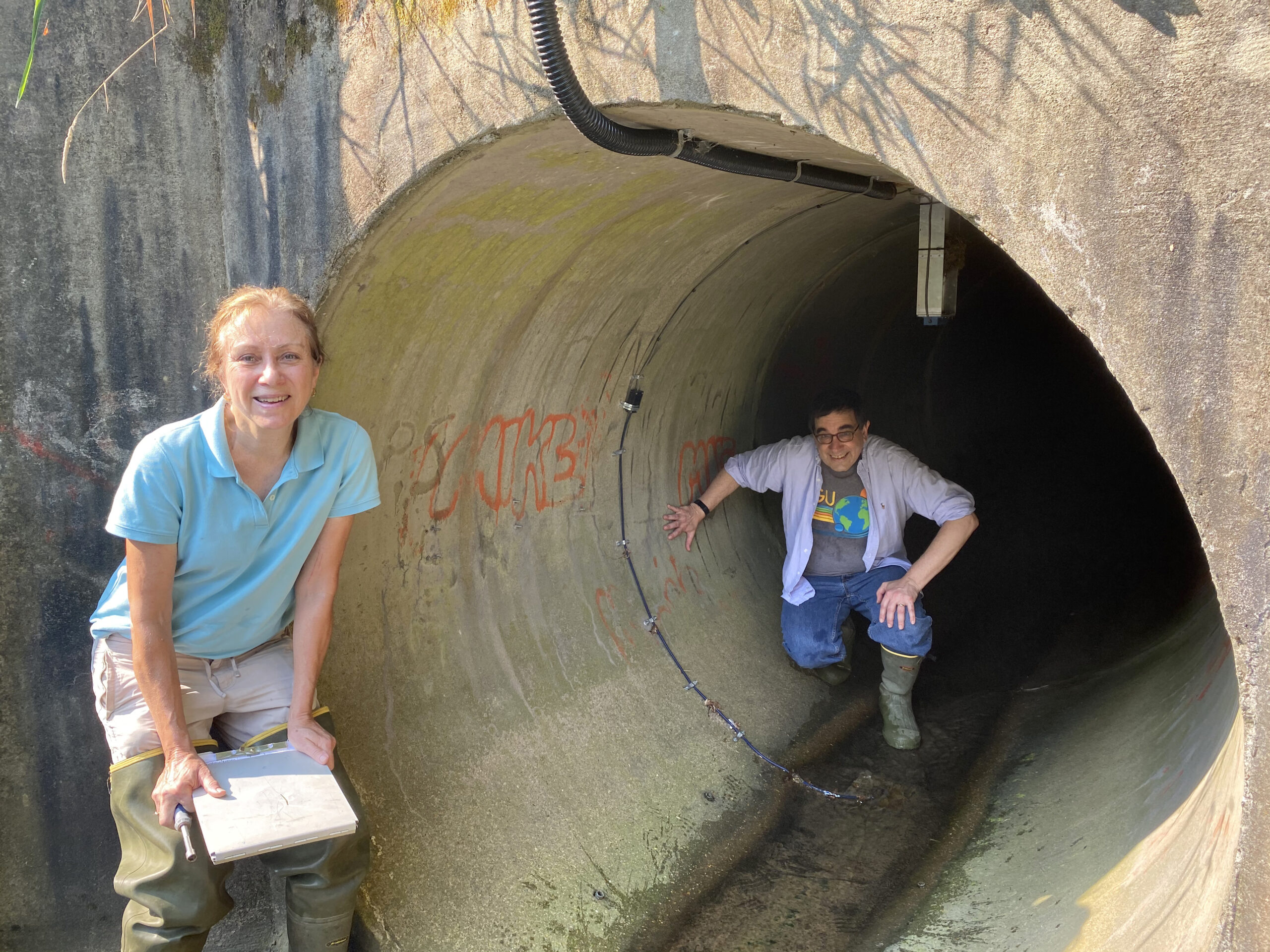 two researchers, one just outside and one inside a large underground pipe several feet in diameter