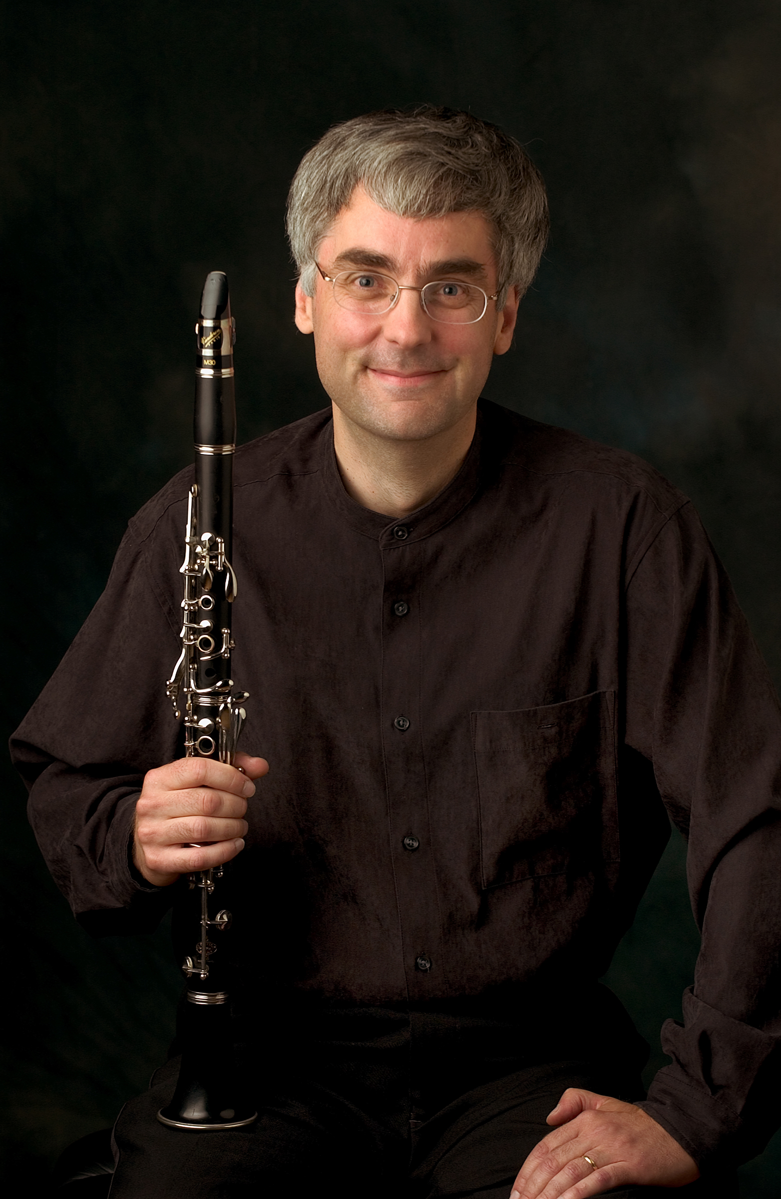 A white man with greying hair, wearing a dark outfit, holds a clarinet