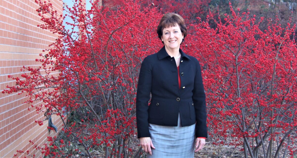 Portrait of Hilltop Institute director, a person with short brown hair wearing a black blazer and grey skirt standing next to a brick building with a red bush in the background.