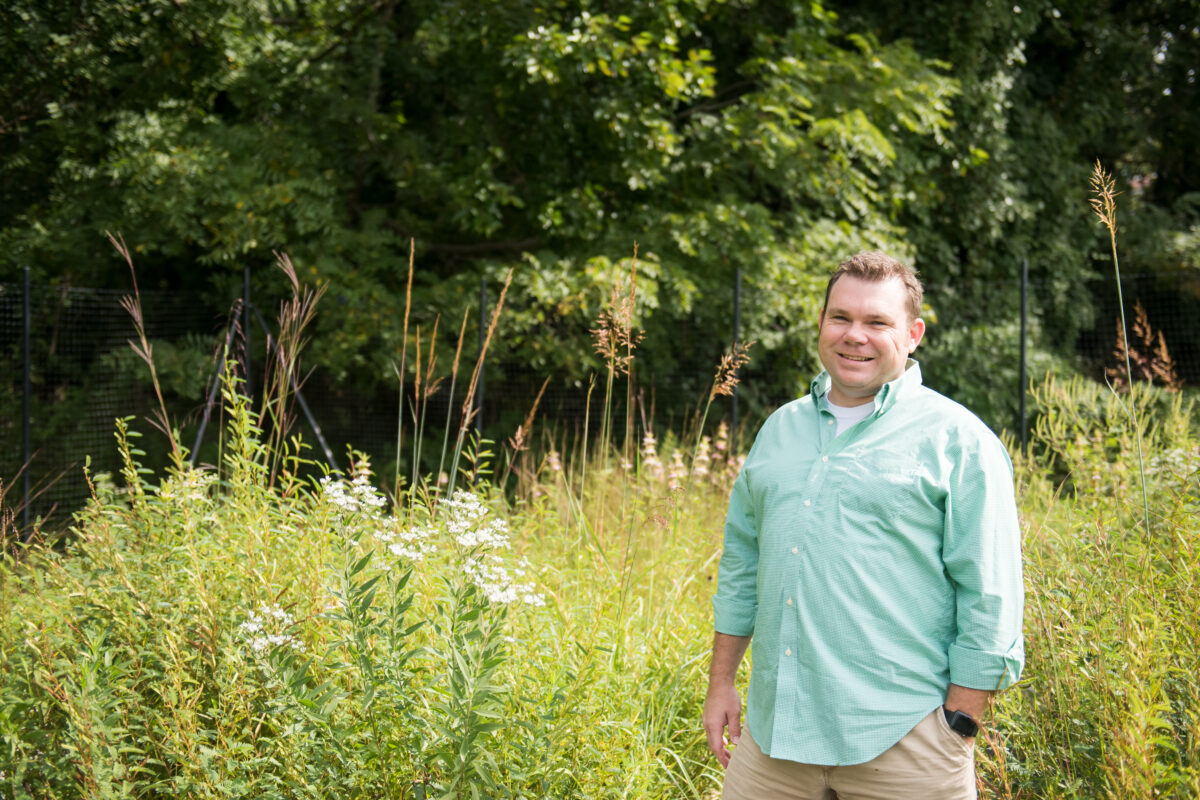 man stands in front of milkweed and other native plants, trees in background
