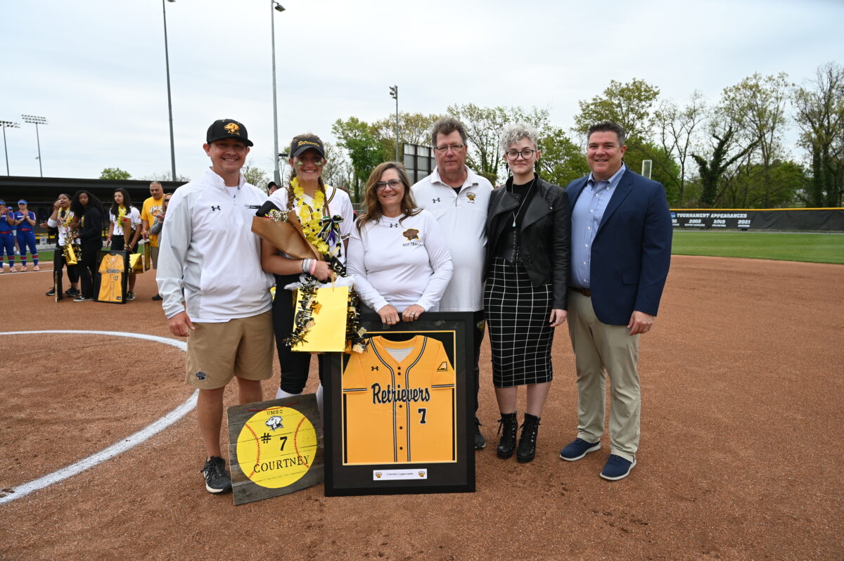 A family, coach, and athletic director stand with a softball player on the softball infield holding a framed jersey
