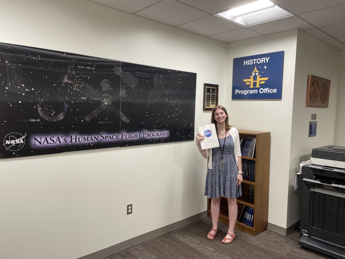 Intern Madelyn Pollack stands next to a NASA poster and holds a NASA binder
