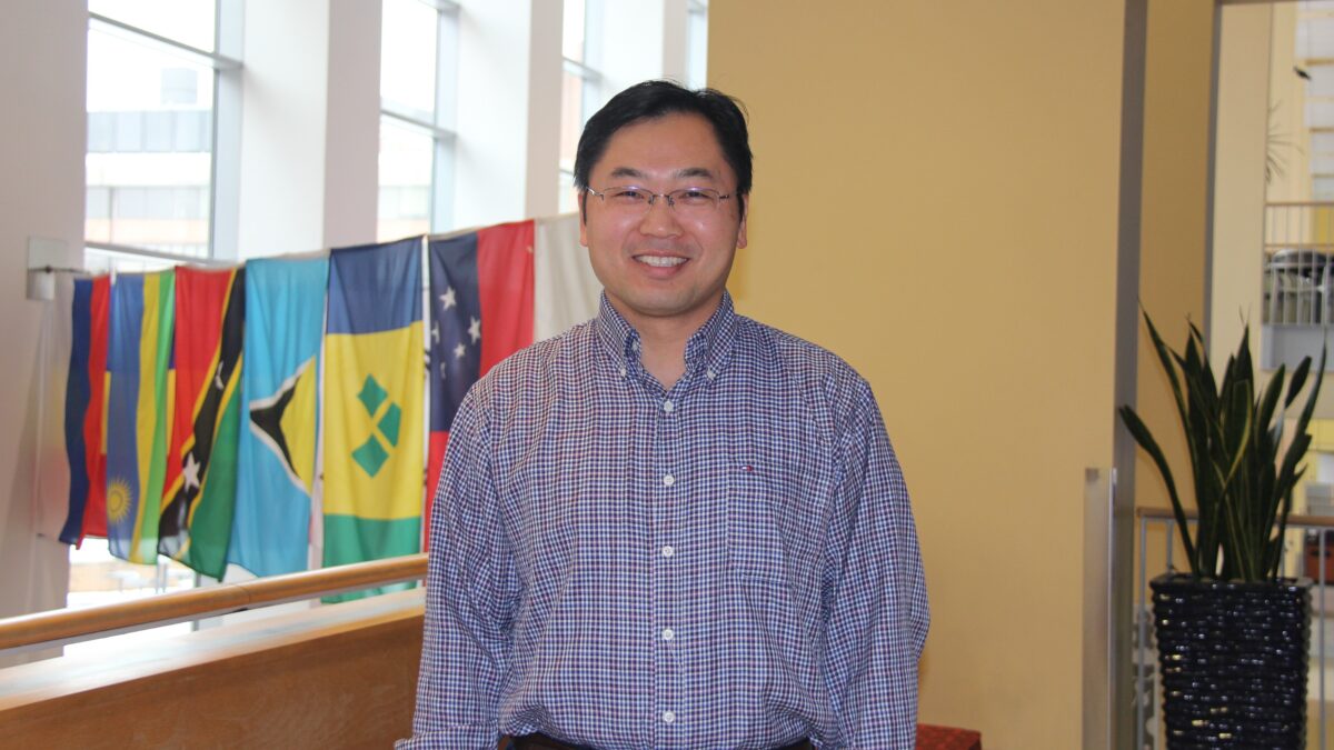 A person with short black hair wearing clear-rimmed eyeglasses and a red and white checkered shirt stands in front of a group of hanging flags from different countries. 