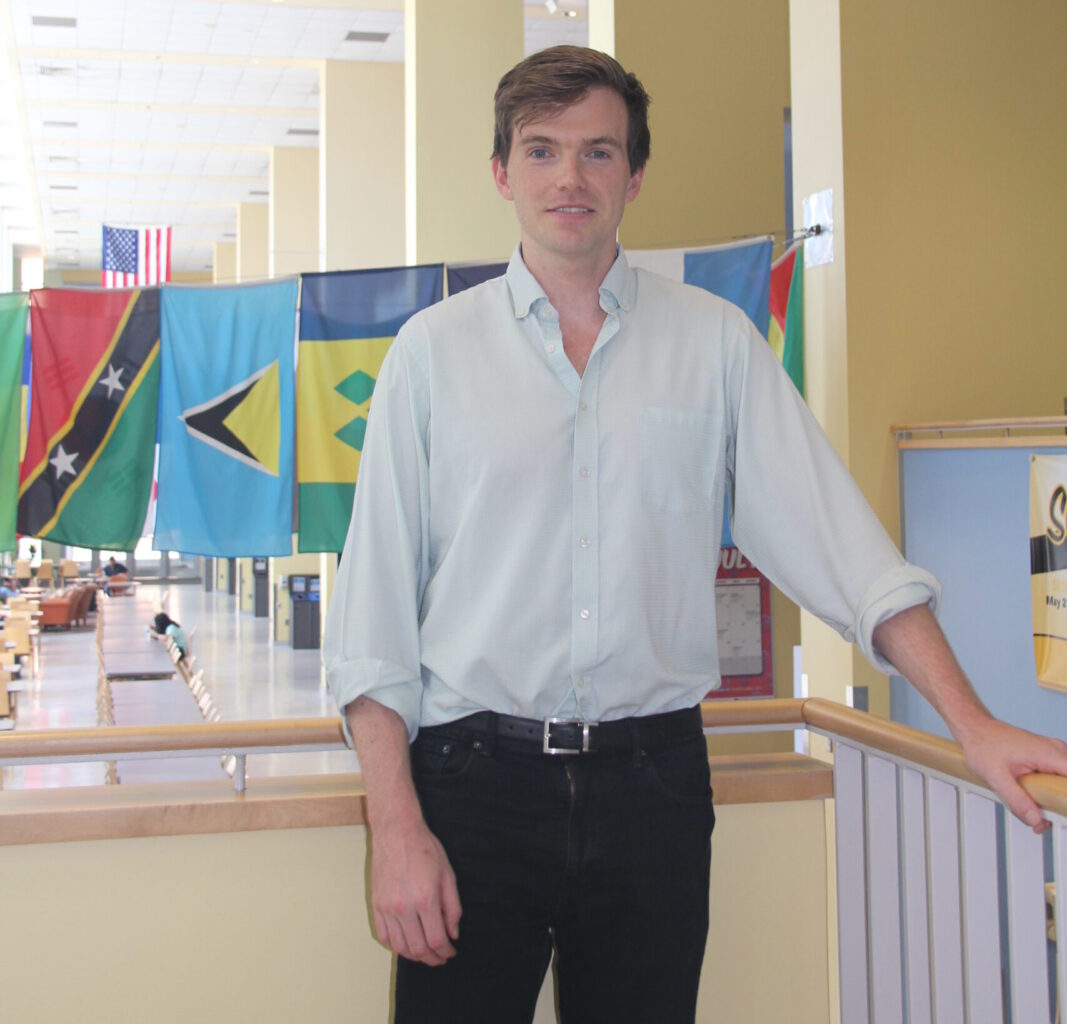 A person with short brown hair wearing a light blue dress shirt with black jeans stands with flags of different countries in the background. Data.