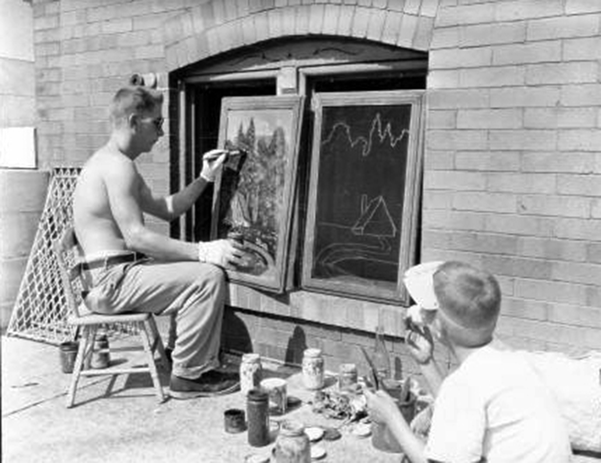 In a black and white photo, a man paints a window screen while a boy looks on