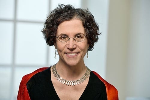 A white woman with glasses is smiling towards the camera. She has on silver jewelry and is wearing a red and black top.
