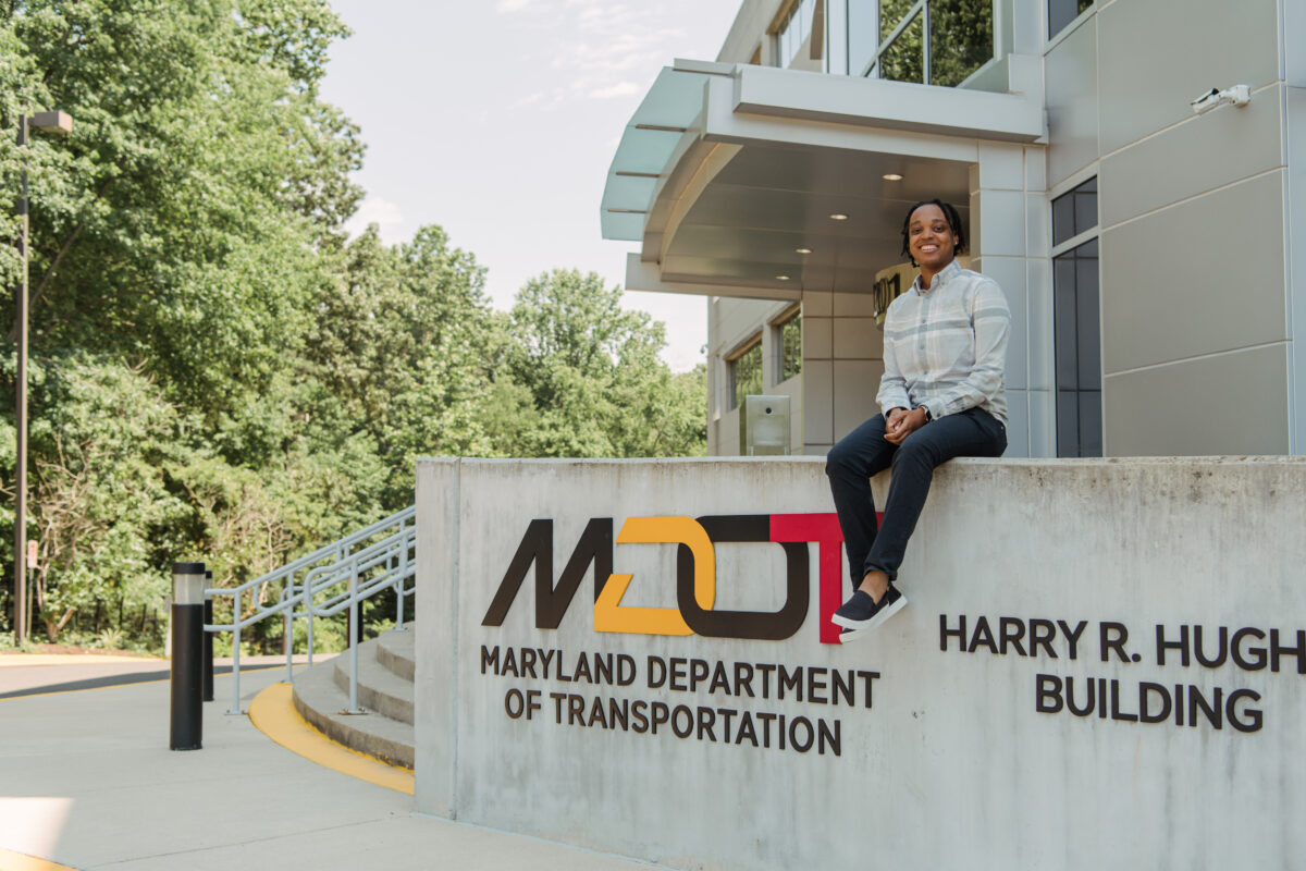 Student (an intern through the Maryland Institute for Innovative Computing) sits on concrete sign in front of an office building. The sign reads "MDOT: Maryland Department of Transportation" and "Harry R. Hughes Building" with the "es" in "Hughes" not visible.