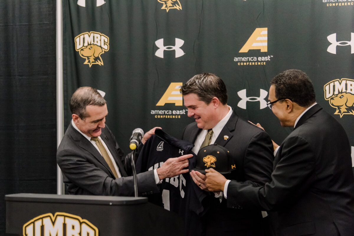 Three men stand around a podium at a press conference. One man holds up a basketball jersey to another man.