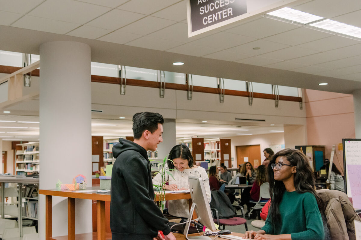 Two students talking at a desk in a library. Sign above them reads "Success Center"