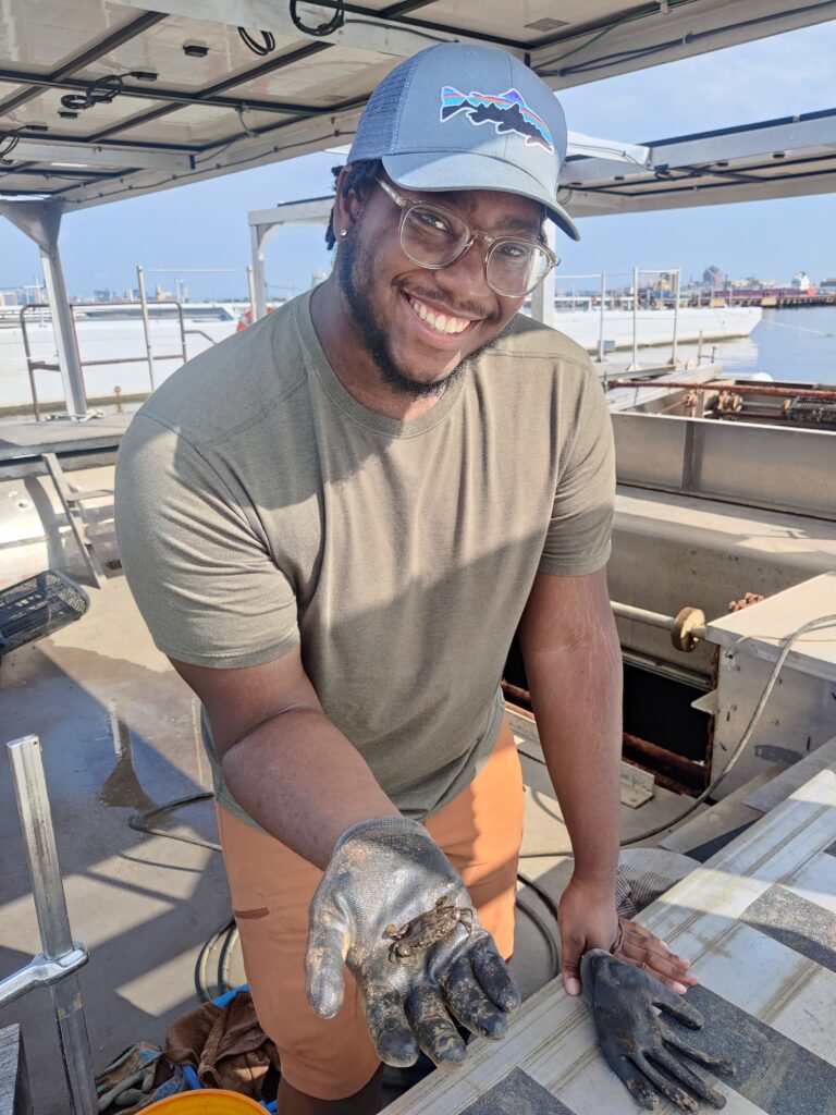 Darryl Acker Carter smiling and holding out a small blue crab in his palm while on a boat