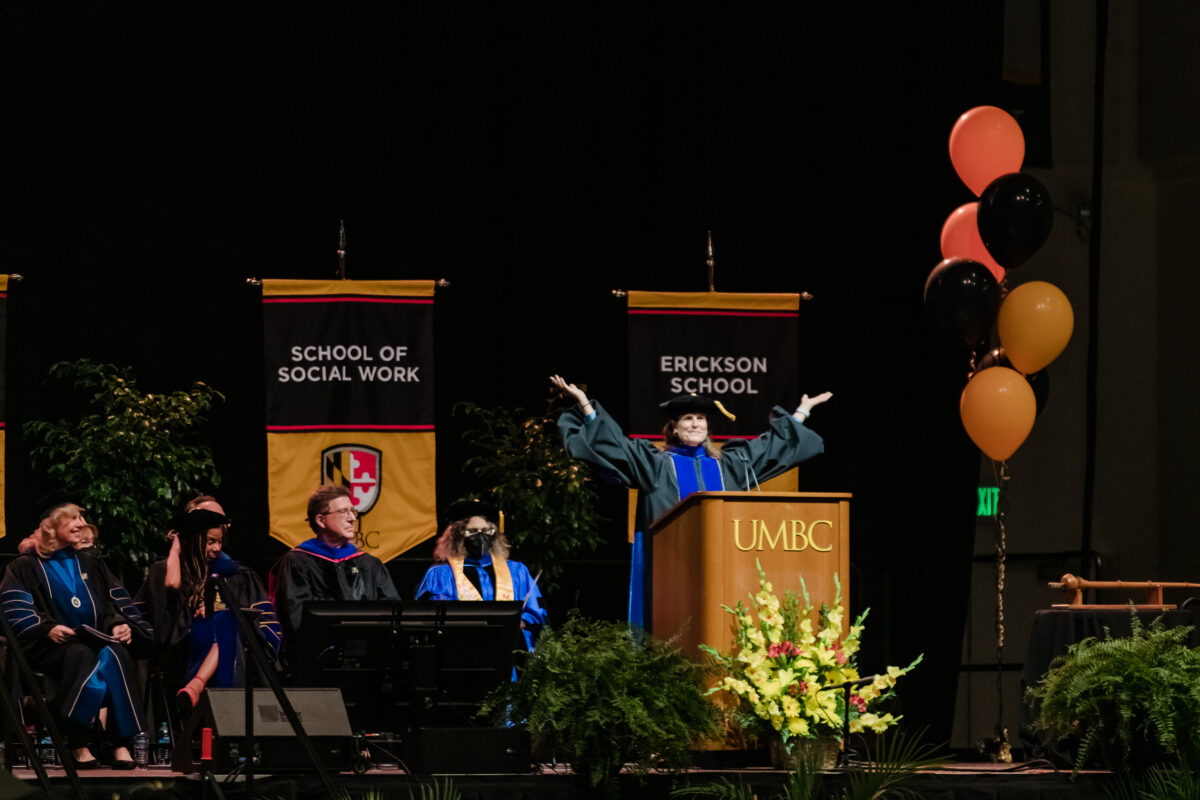 Woman in university regalia at podium with arms outstretched smiling