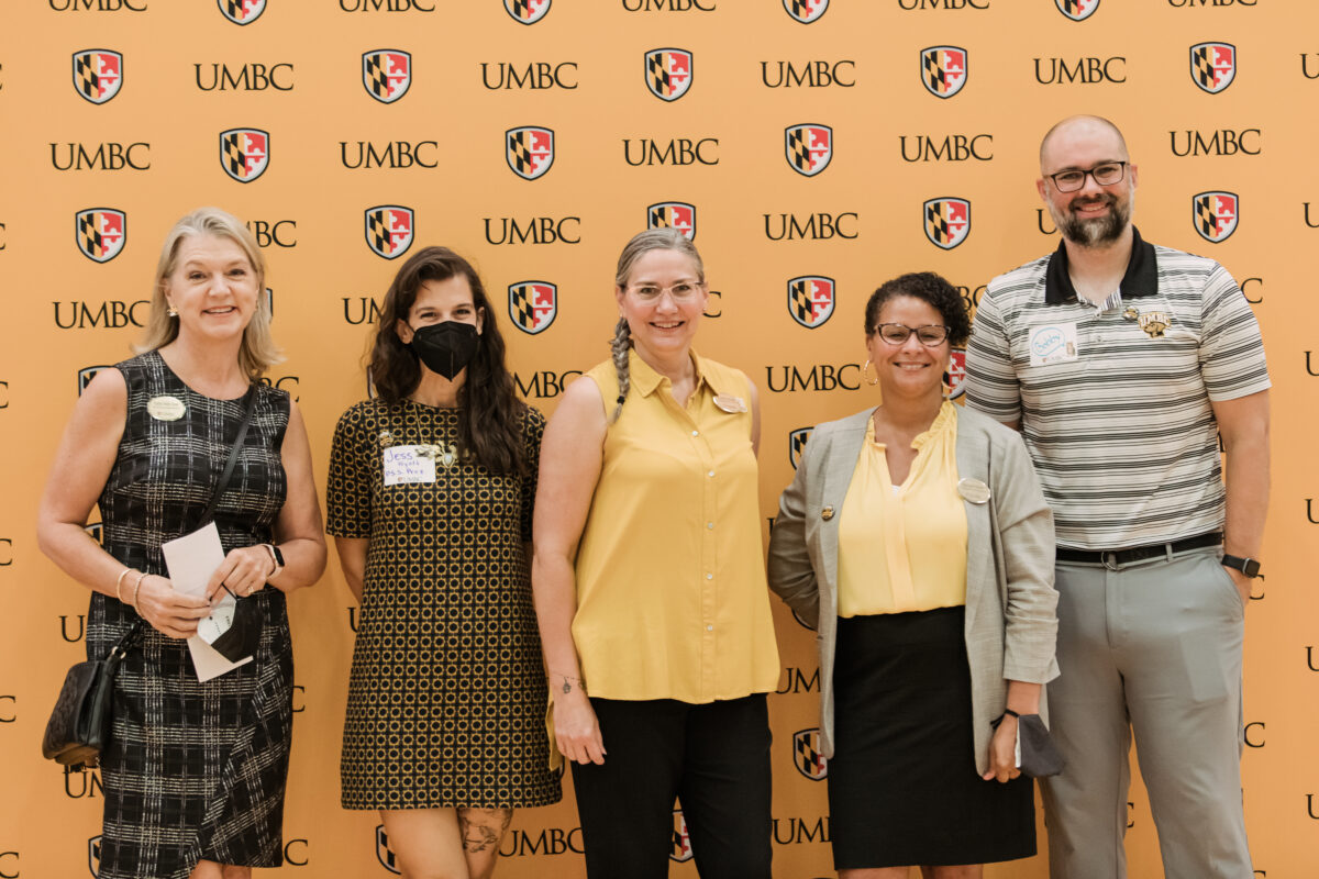 Five adults wearing professional clothing in different black and gold patterns smile in front of a sign reading "UMBC" several times.