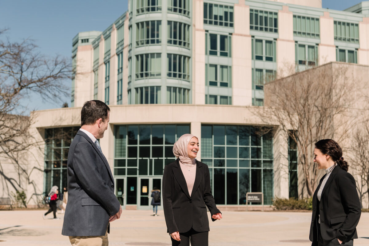 Three people in professional clothing talk in front of a large glass and concrete building