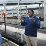 Darryl Acker-Carter speaking on a dock, with the floating oyster aquaculture setup behind him, to a group of teachers