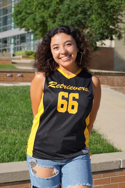 A woman with curly brown hair and in a Retrievers 66 jersey stands in front of green grass