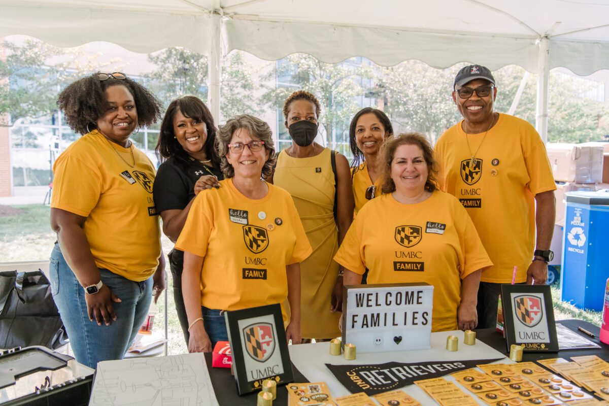 A group of parent volunteers in yellow shirts smile for a camera with "Welcome families" signs