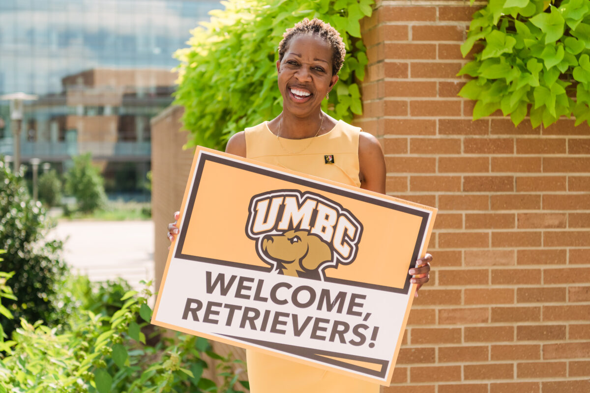 Smiling woman in gold dress stands outside holding a sign that reads, "Welcome, Retrievers!"