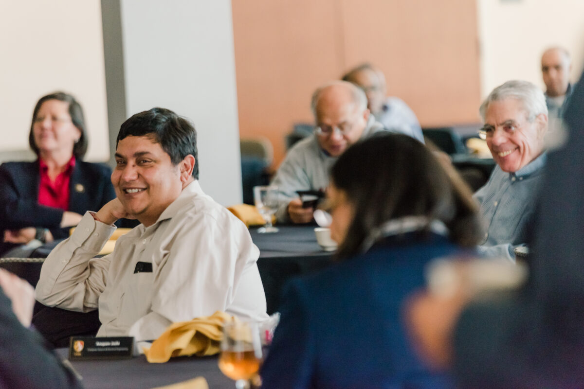 Several people in professional attire sit at tables in a room. Focus is on one man who is smiling.