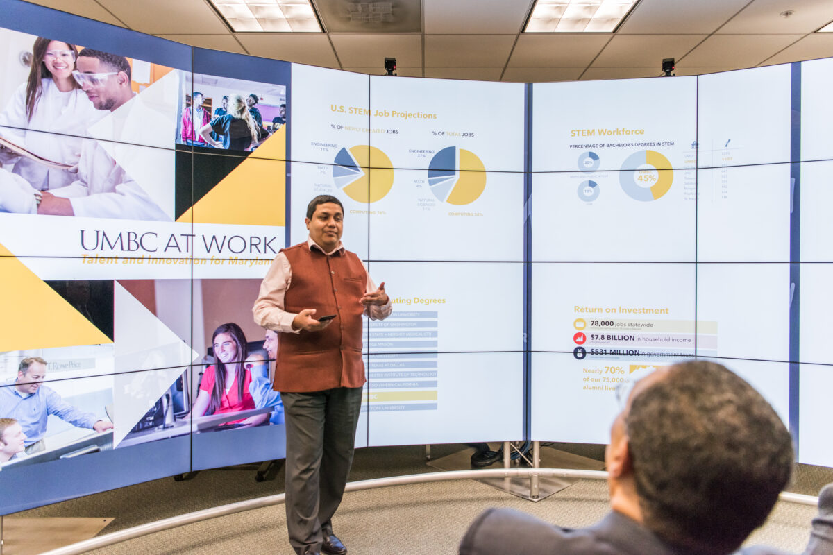 Man in professional attire (Anupam Joshi) presents information that is shown on a very large screen behind him. The screen reads, "UMBC at Work."