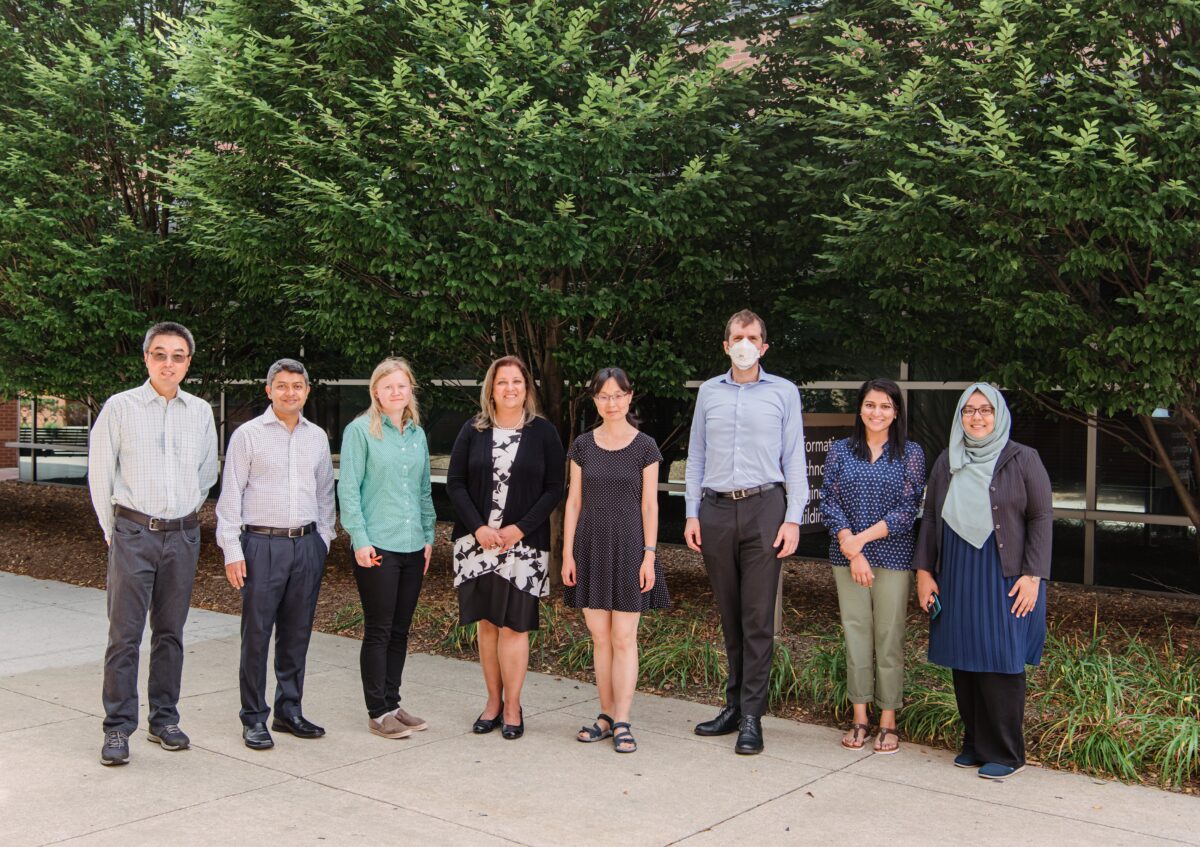 A group of eight people in professional attire stands for a portrait in front of a building and trees.