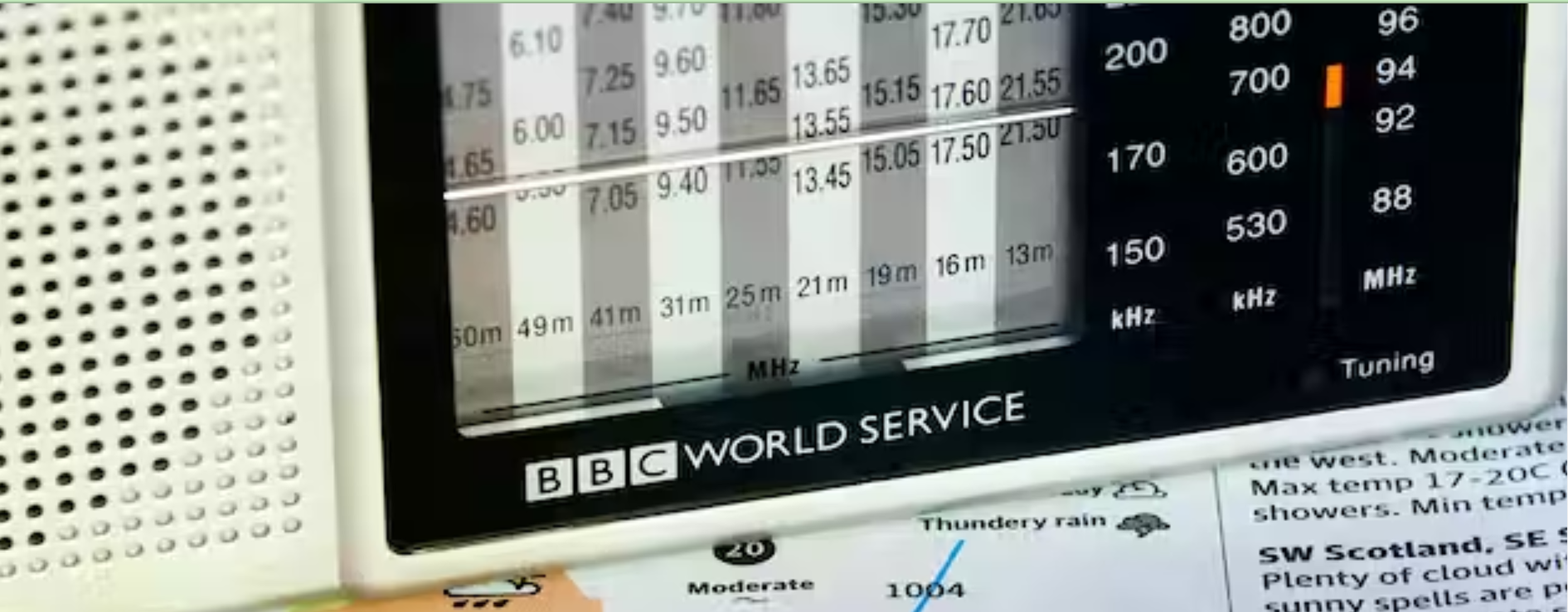 a black and white transistor radio with the words BBC World Service in white lettering sits on top of a a newspaper BBC