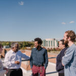 five people stand on a rooftop with a blue sky and the UMBC library in the background