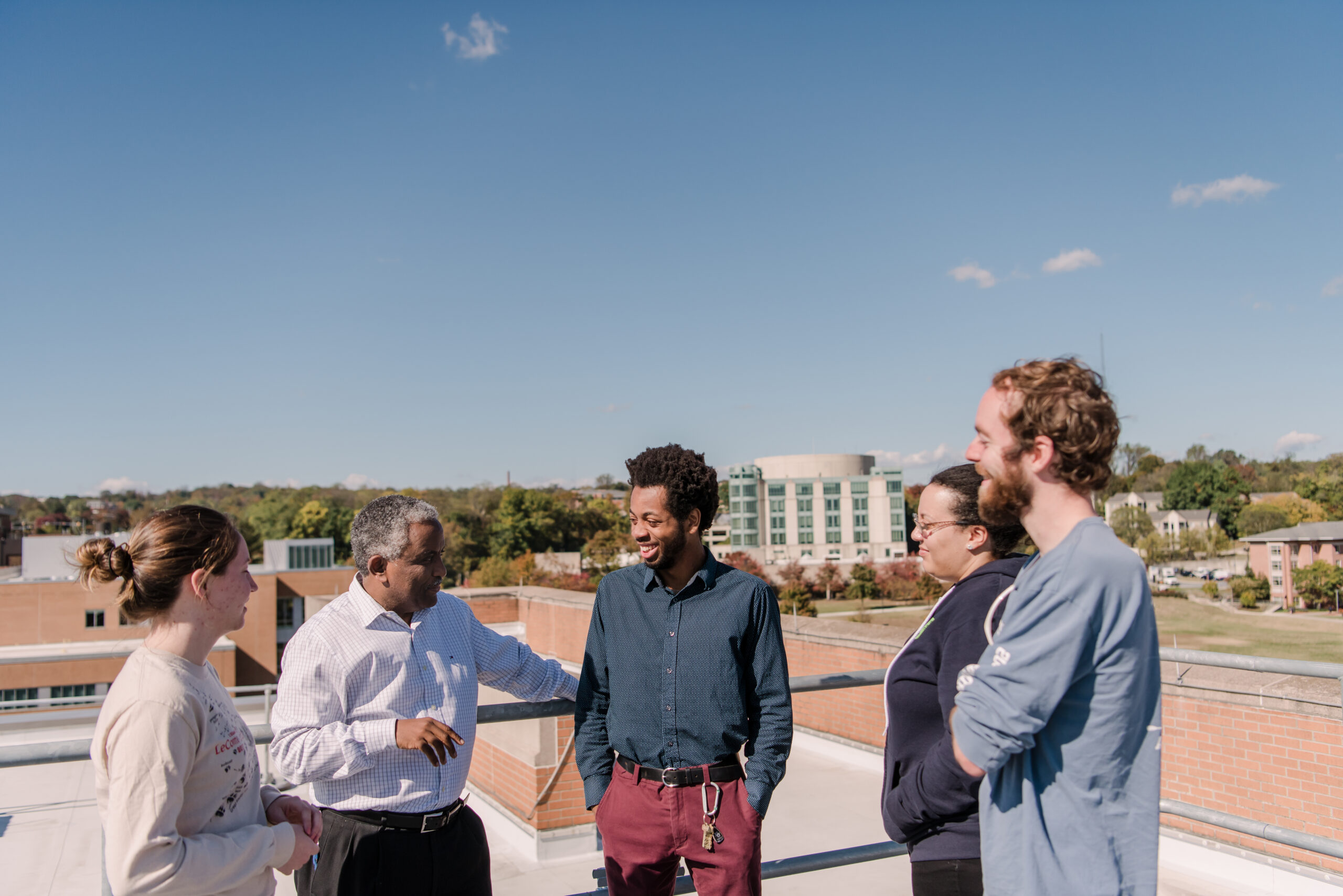 five people stand on a rooftop with a blue sky and the UMBC library in the background