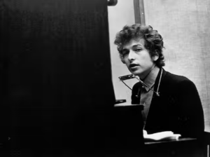 A person sits at an upright piano with a harmonica strapped around their neck.