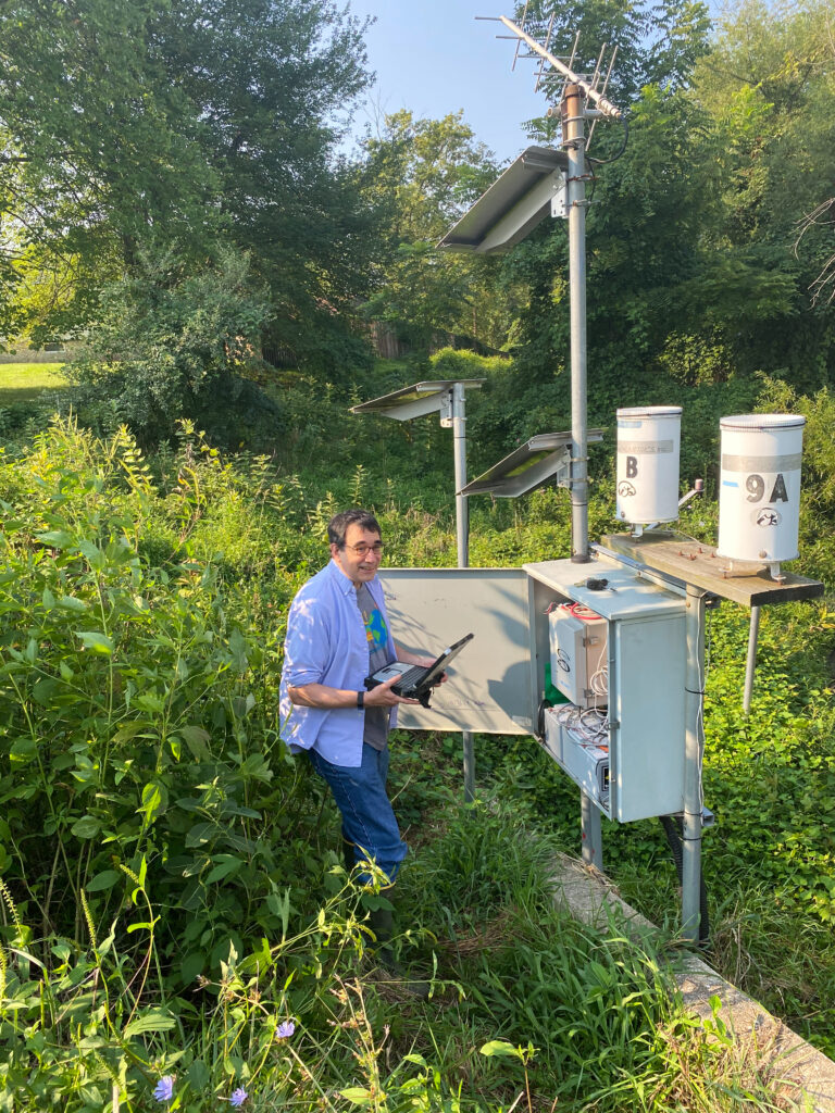 A researcher standing among greenery and holding a clipboard looking at a sampling station, which looks like an transformer box with the frontn panel open, with a small solar panel attached to it on a tall pole 