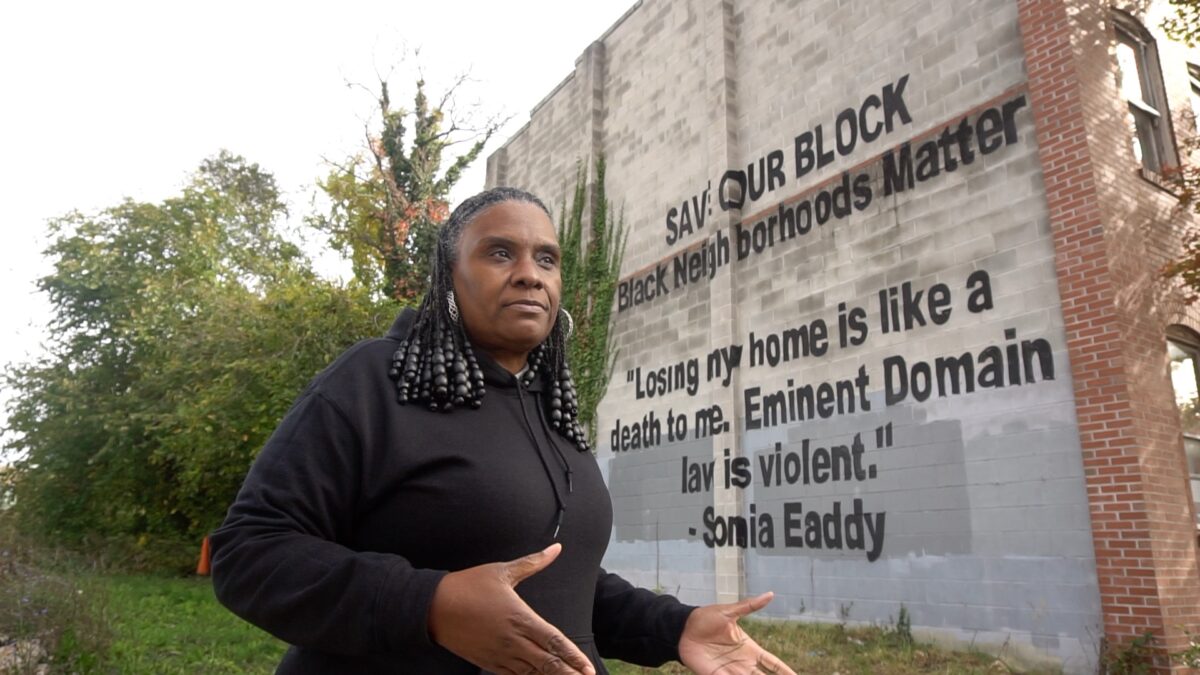 A person with long black hair and wearing a black hoodie stands in front of a brick wall with a mural of a quote with large black letters. Public humanities. Poppleton Baltimore.