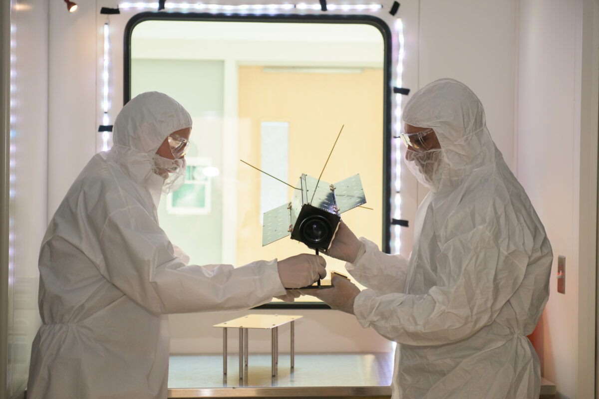 two researchers in all-white, full-body protective gear hold the HARP cubesat inside a "clean room" laboratory.
