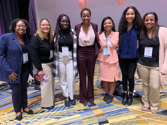 Seven women wearing professional clothing stand together at a conference