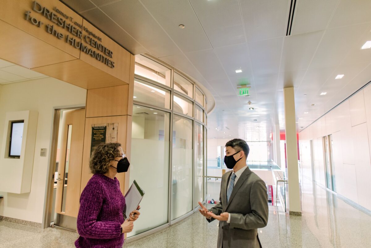 Two people stand inside a well lit building next to a curved glass wall while facing each other. 