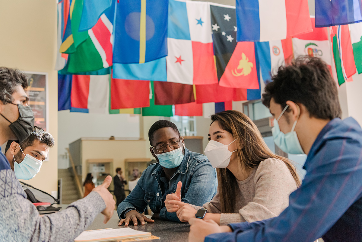 Five masked students sitting and talking beneath a ceiling of hanging flags from different countries.