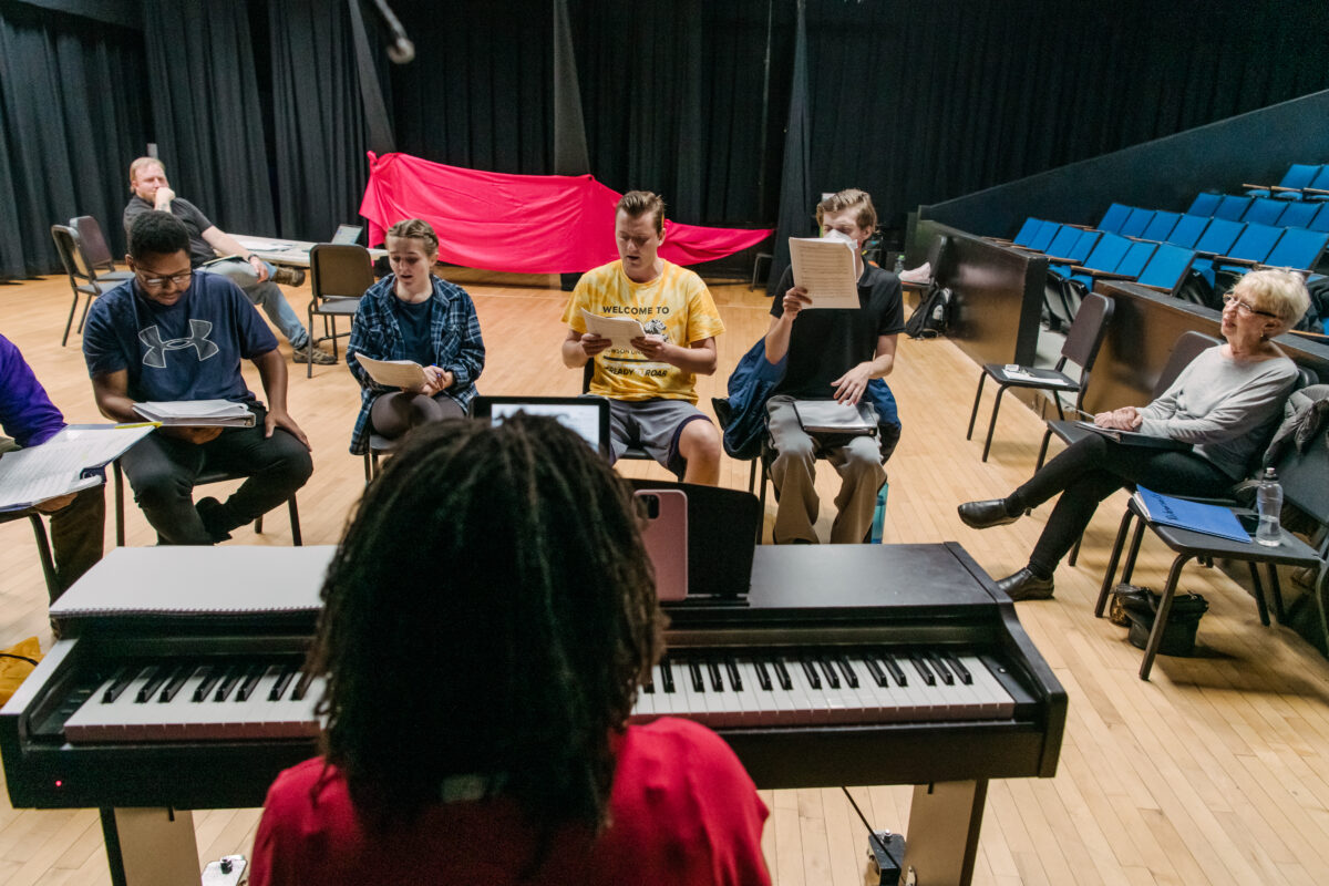 A group of performers sit around a piano.