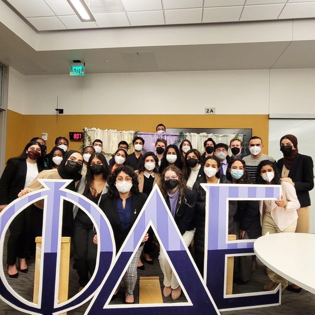 Members of the medical fraternity Phi Delta Epsilon pose together in front of a large sign of their greek letter.