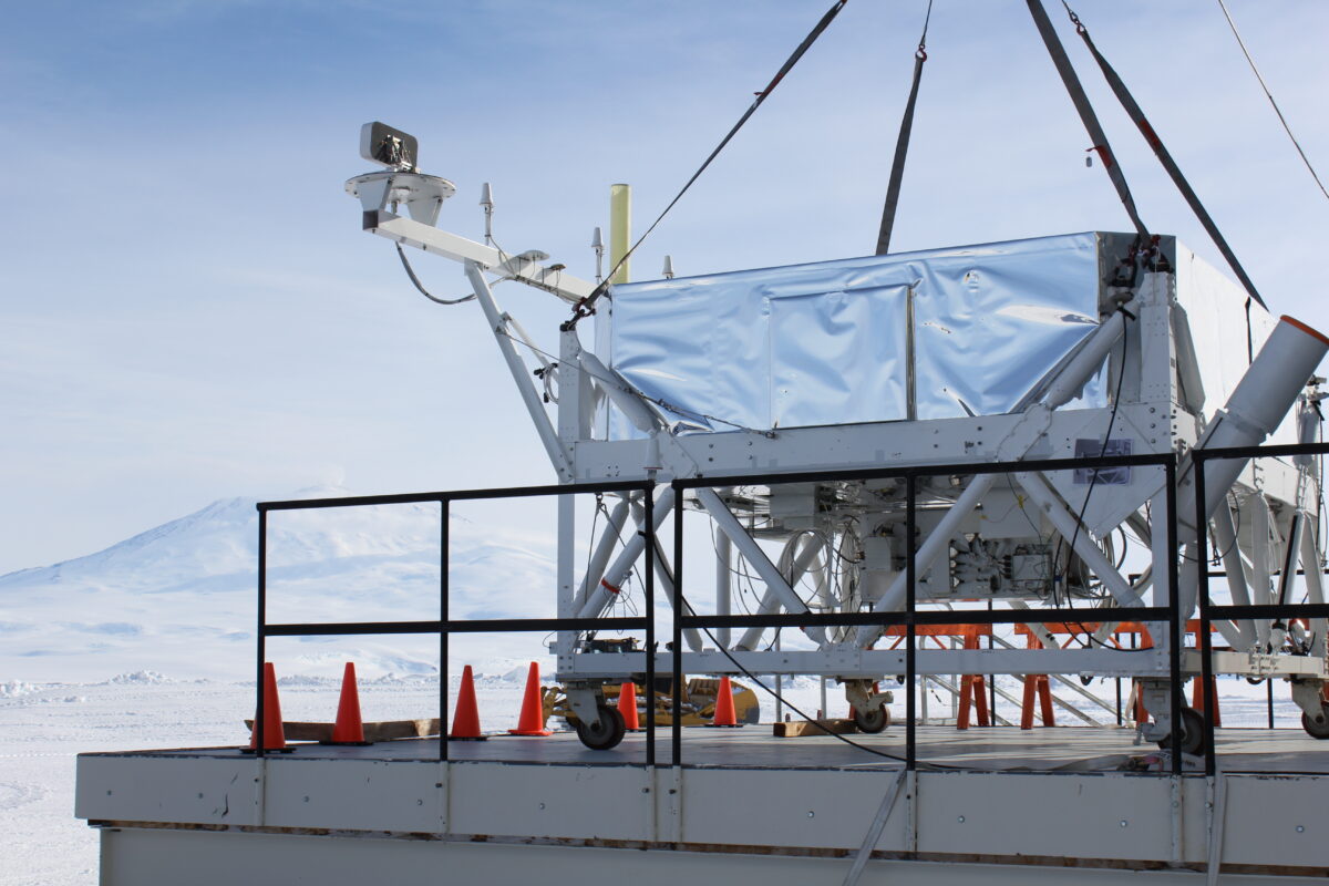 Snowcapped peaks in the background. A metal box about the size of a shipping container rests on a platform, suspended from above by large cables, surrounded by orange cones on the ground.