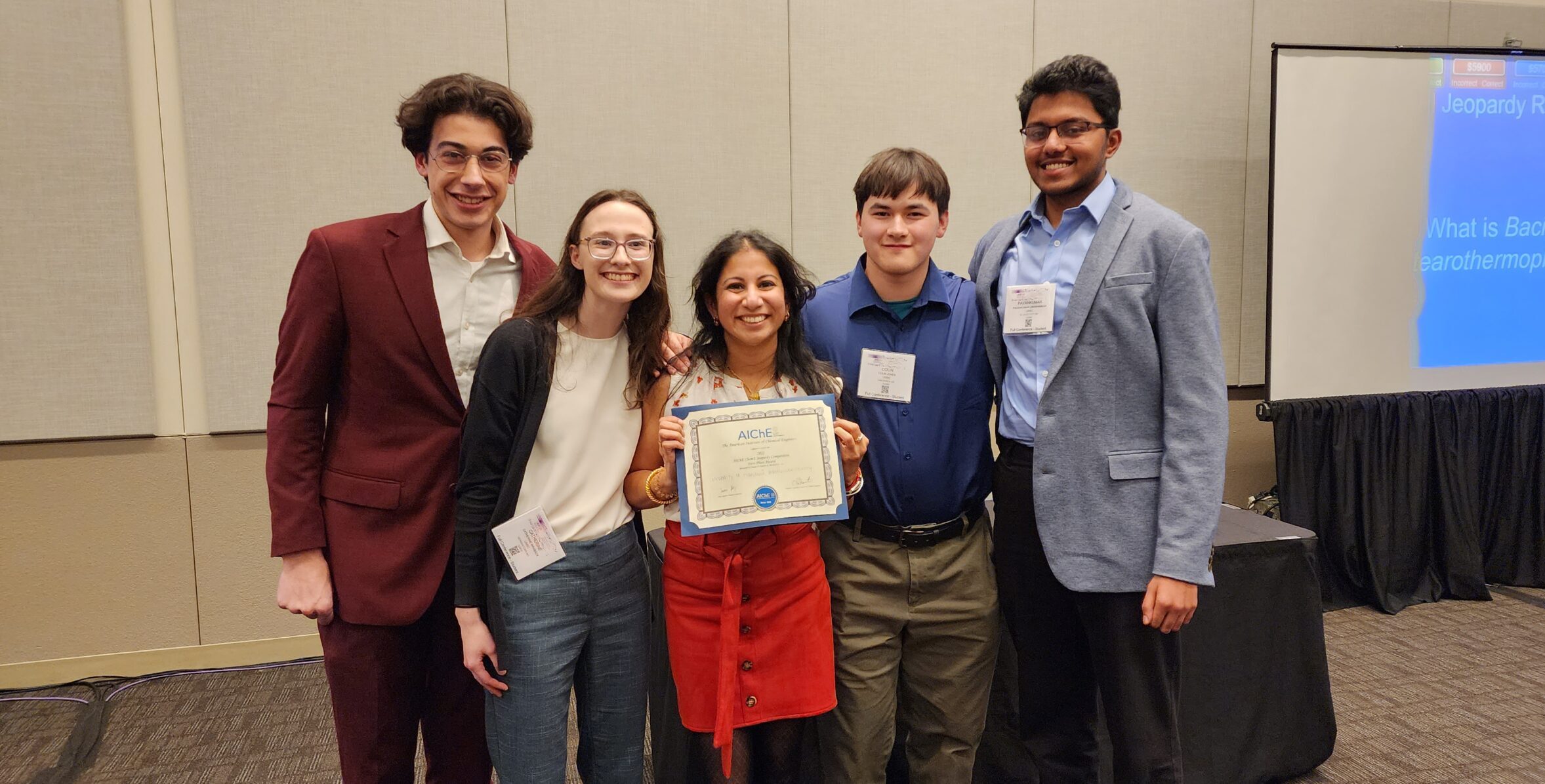 Five smiling people in professional clothing pose for a portrait in a conference room with one holding an award certificate reading AIChE ChemE Jeopardy Competition First Place.