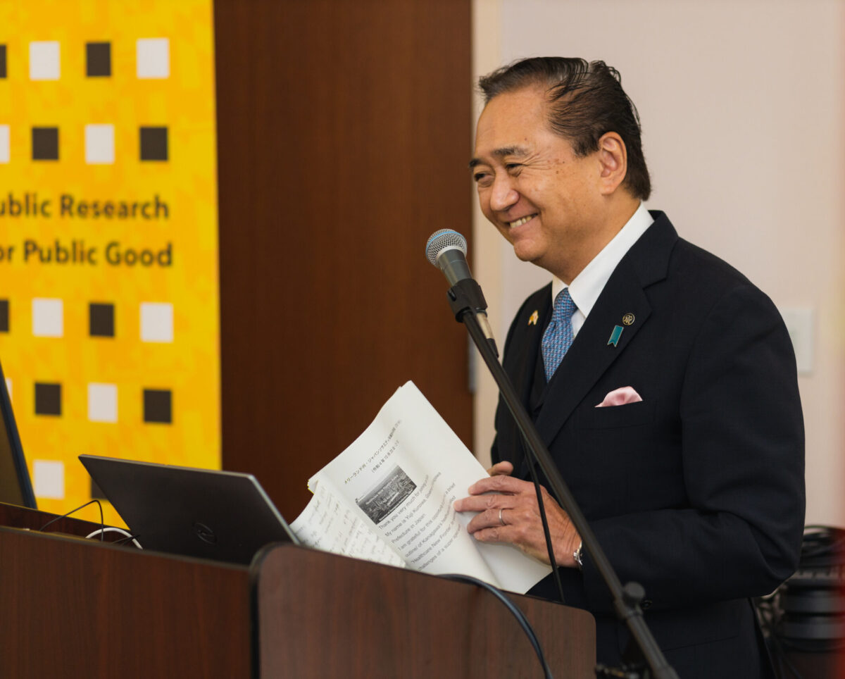 A person with a black suit stands smiling at a podium with a laptop and a microphone holding white sheets of paper with black writing and a gold, white, and black banner in the background with the words "Public Research for Public Good"
