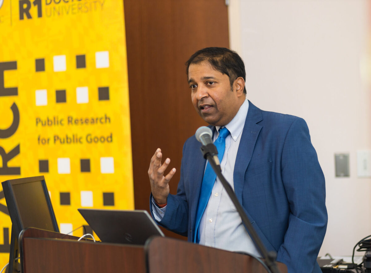 A person in a blue suit stands at a podium with a laptop and a microphone with a gold, black, and white banner with the words UMBC R1 Doctoral University.