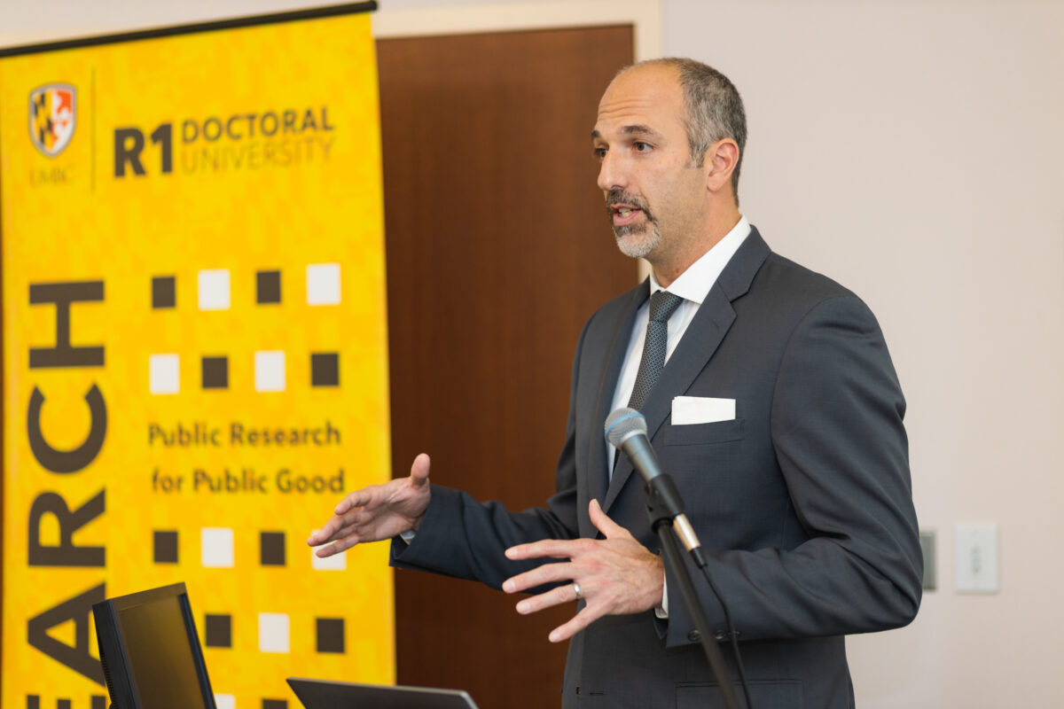 A person wearing a dark grey suit stands at a podium with a laptop and a microphone and a gold, black, and white banner in the background with the words UMBC R1 Doctoral University 