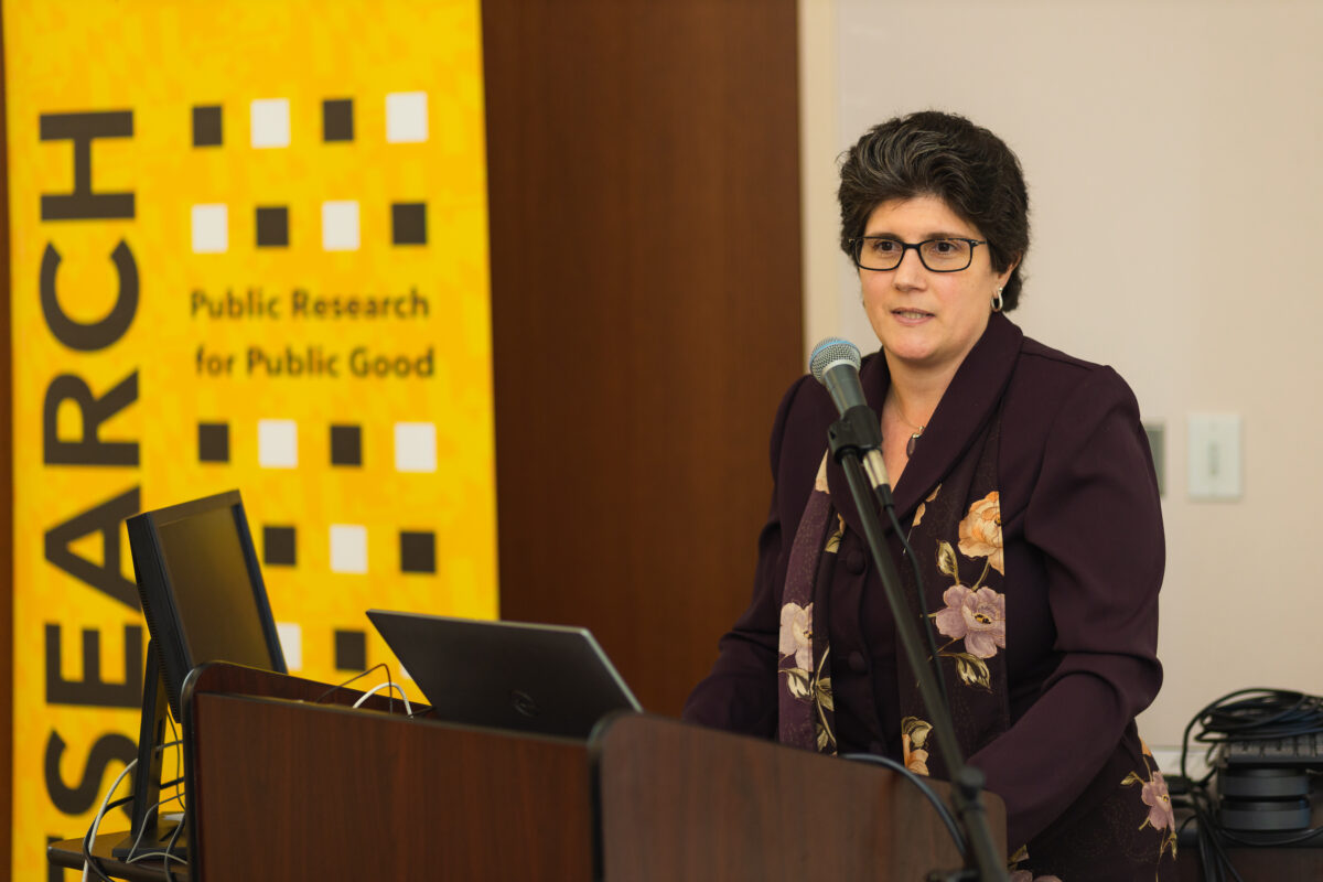 A person wearing a plum dress coate with a scarf with purple and yellow flowers stands at a podium with a laptop and a microphone with a gold, black, and white banner with the words UMBC R1 Doctoral University.