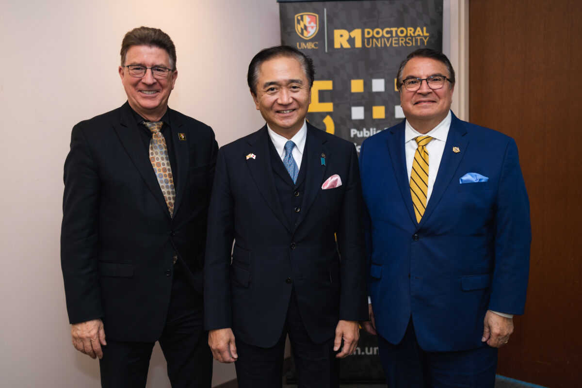 Three adults wearing business suits stand side by side smiling at the camera with a black and gold banner in the background with the words UMBC R1 Doctoral University.